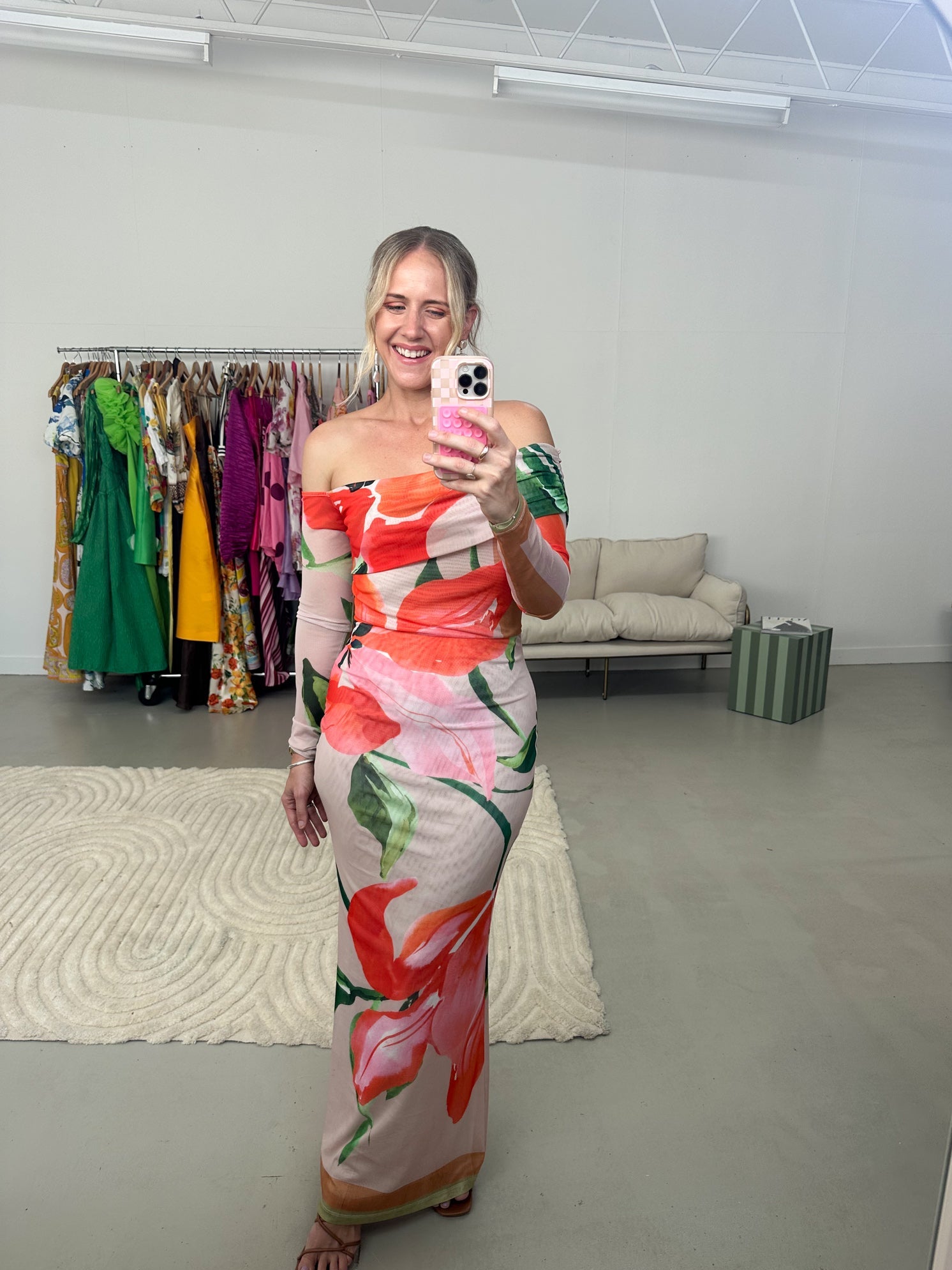Woman taking a mirror selfie wearing a floral off-shoulder dress in a clothing store.